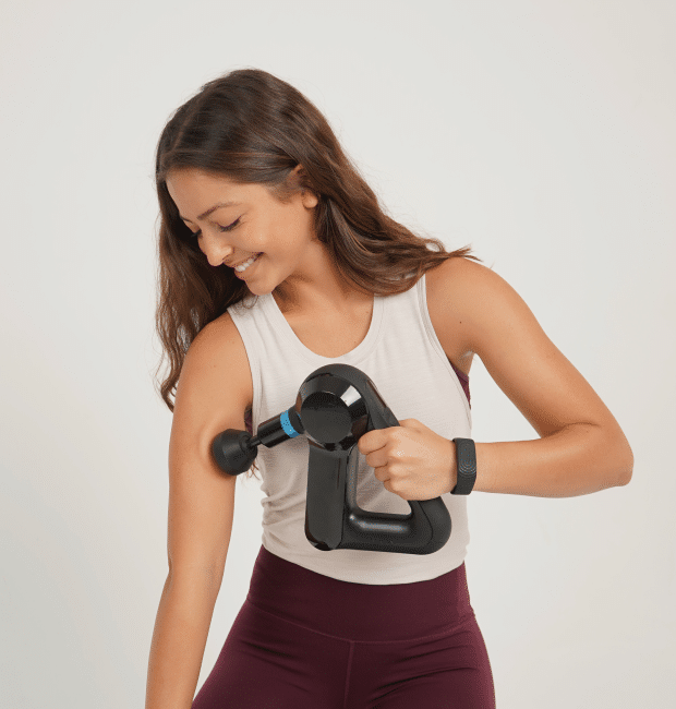 A woman in workout attire uses a handheld percussive massage device on her upper arm, promoting stress resilience while supporting ANS balance.