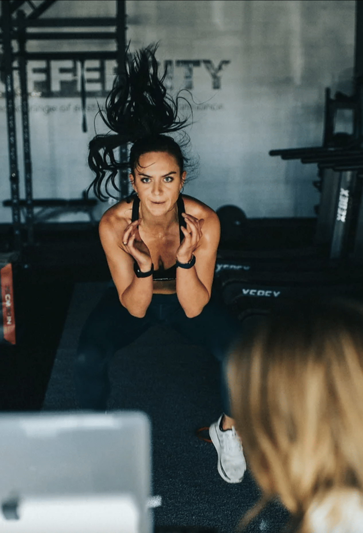A fit woman with long ponytail hair is captured mid-jump while exercising intensely at the gym. Visible gym equipment sets the scene in the background, with another person partially visible in the foreground.